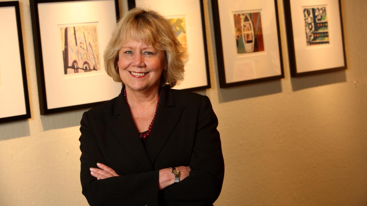 Director Judy L. Larson standing in front of framed artworks, smiling with her arms crossed.