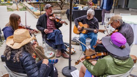 Musicians sitting in a circle outdoors playing lively music on violins and a guitar.